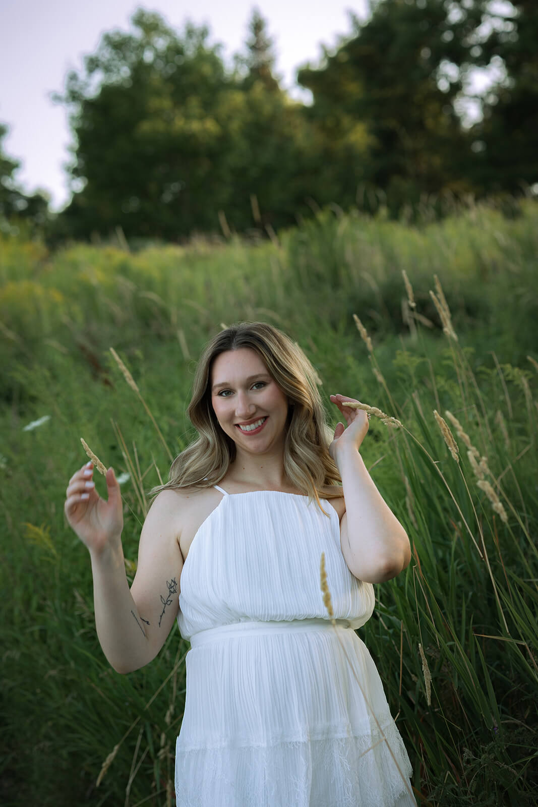 Woman in a white tiered dress smiling at the camera while standing among tall green grass and lifting a hand toward her hair.
