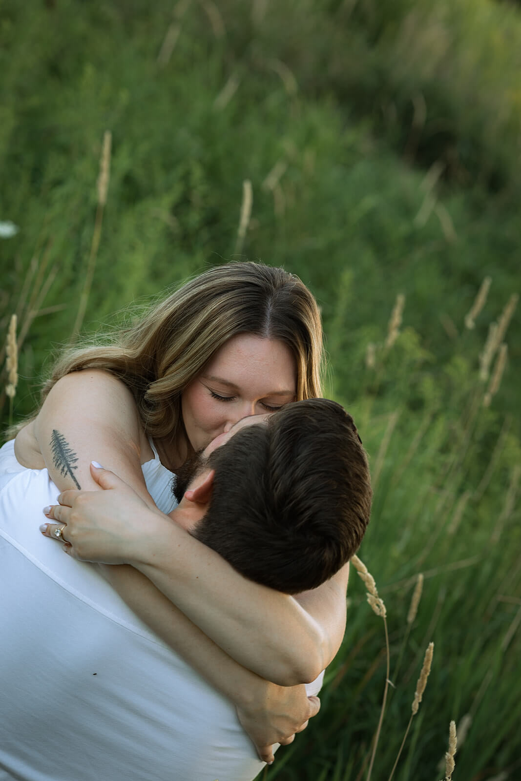 Woman wrapping her arms around her partner and kissing the top of his head while they embrace in tall grass.