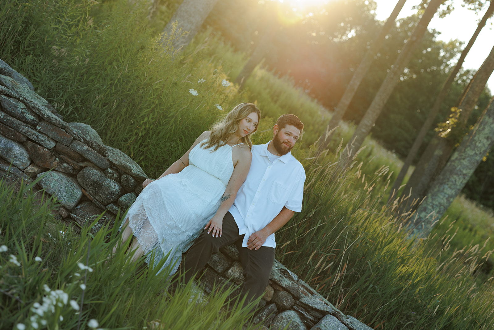 Couple sitting together on a stone wall surrounded by tall grass in the warm evening light at Beech Hill Barn.