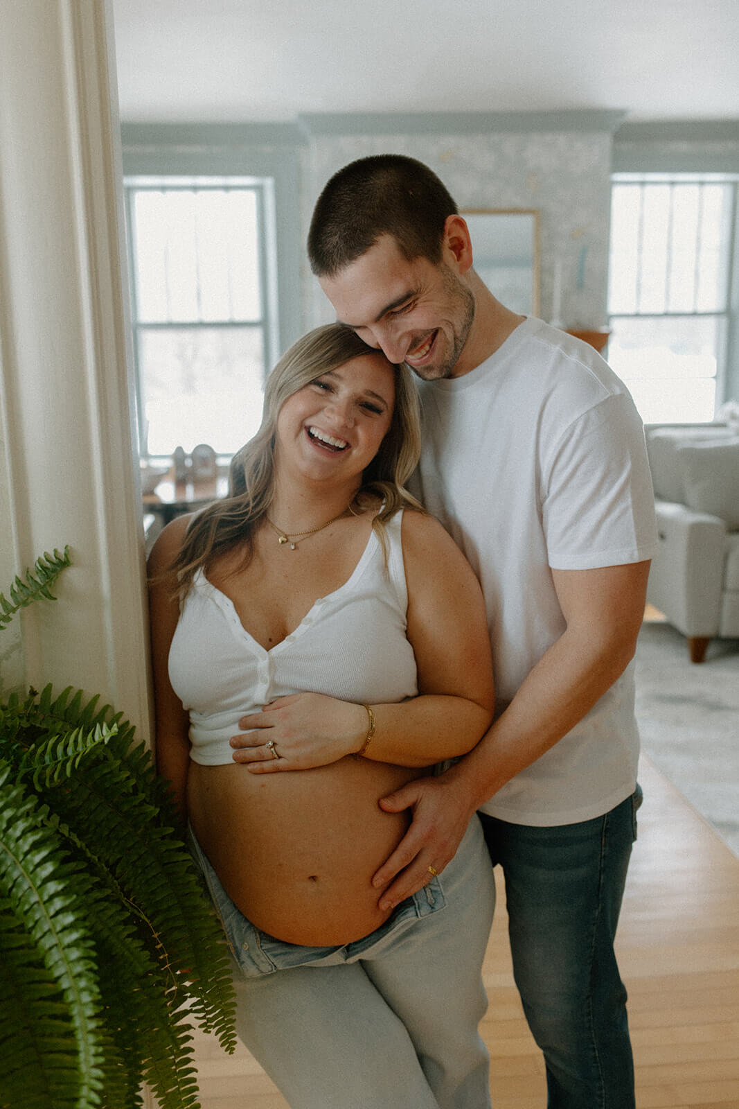 Maternity photoshoot at home of an expecting couple laughing together while he stands behind her with his hands on her belly.
