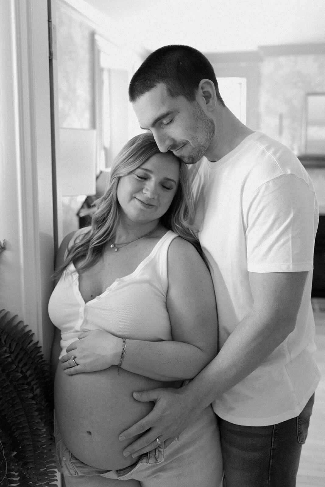 Black-and-white maternity photoshoot at home of an expecting couple embracing with their hands resting on her baby bump.