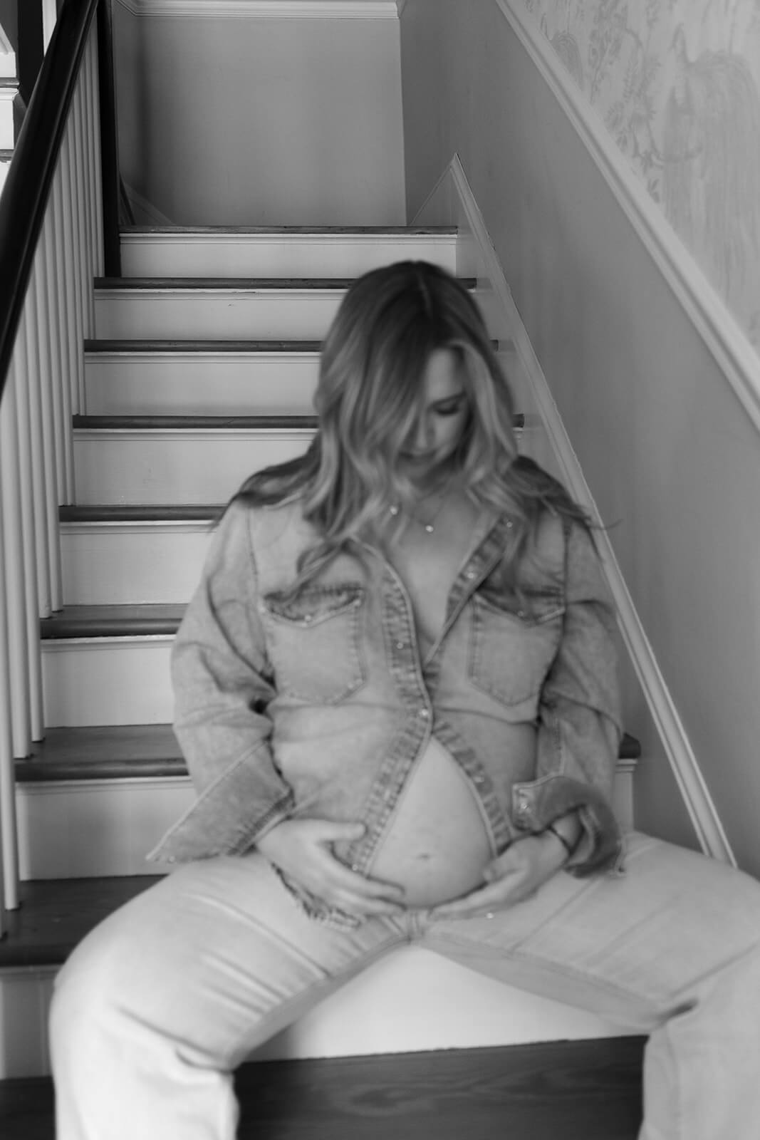 Black-and-white portrait of an expecting mother sitting on the staircase in an open denim shirt.