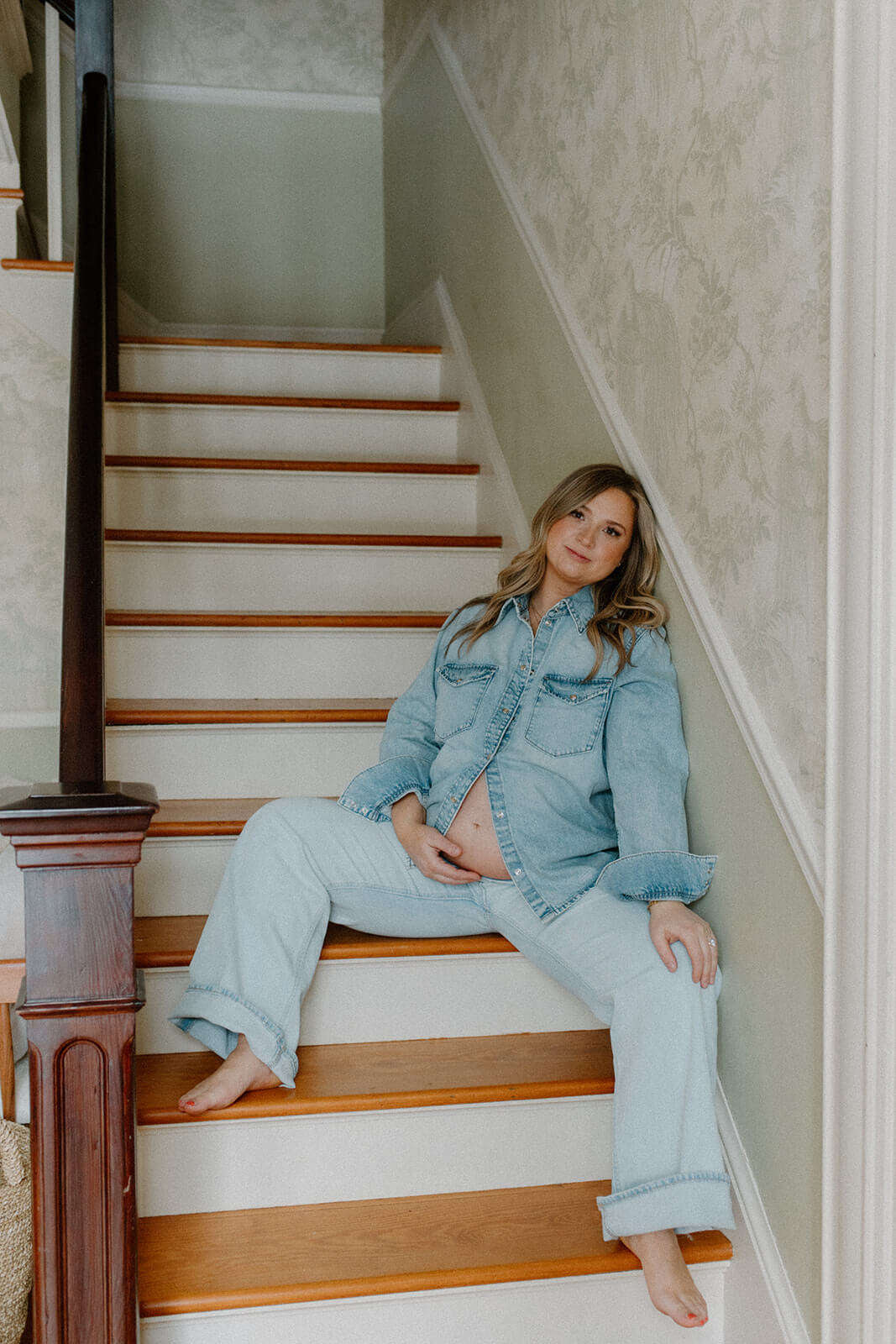 Portrait of an expecting mother sitting on the staircase in a denim outfit with her belly showing.