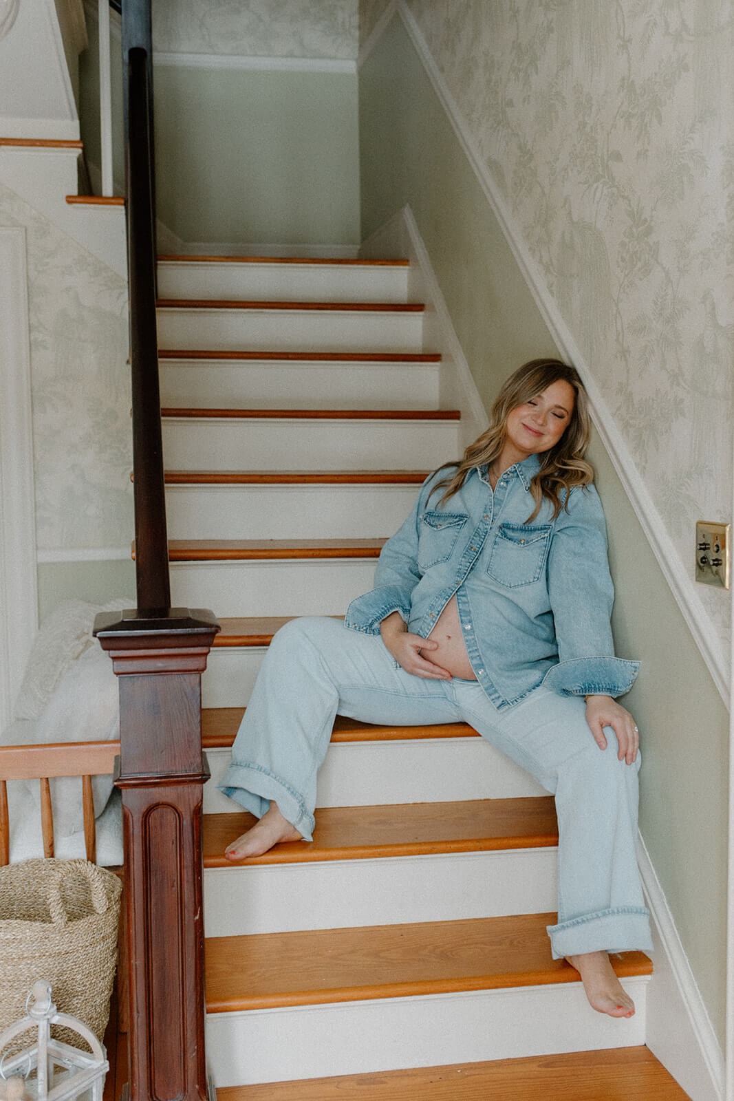 Maternity photoshoot at home portrait of an expecting mother sitting barefoot on the staircase in a denim outfit.