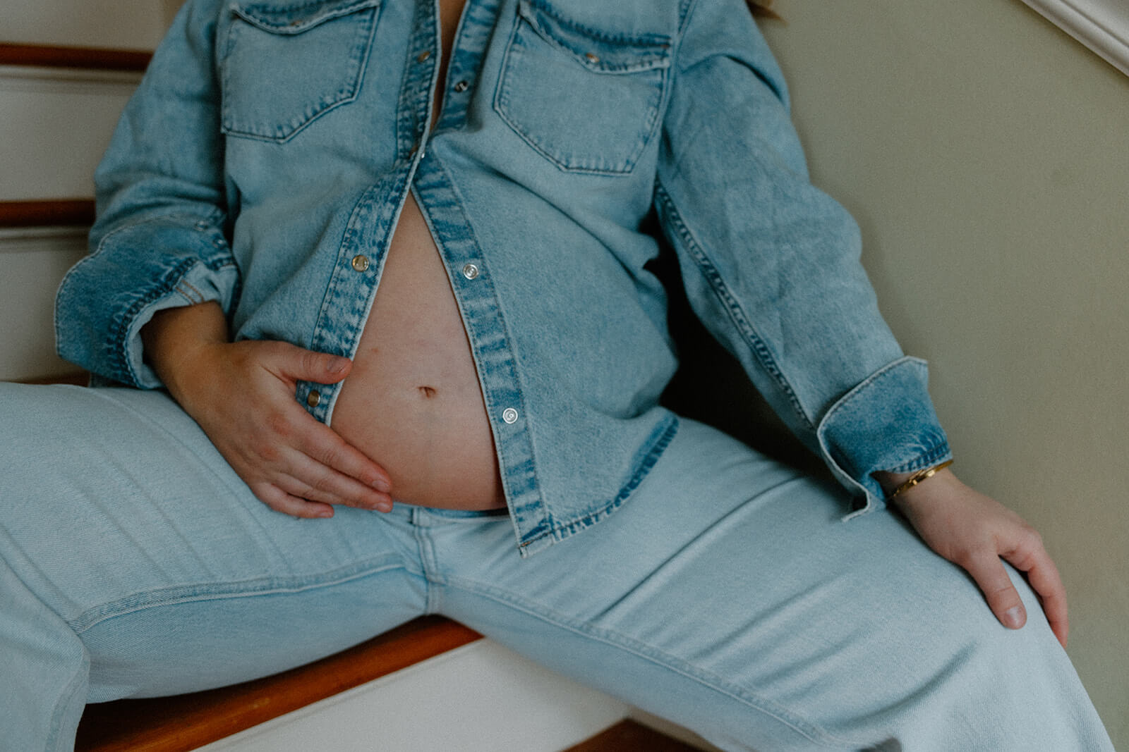 Close-up showing an expecting mother’s bare belly beneath an open denim shirt while sitting on the stairs.