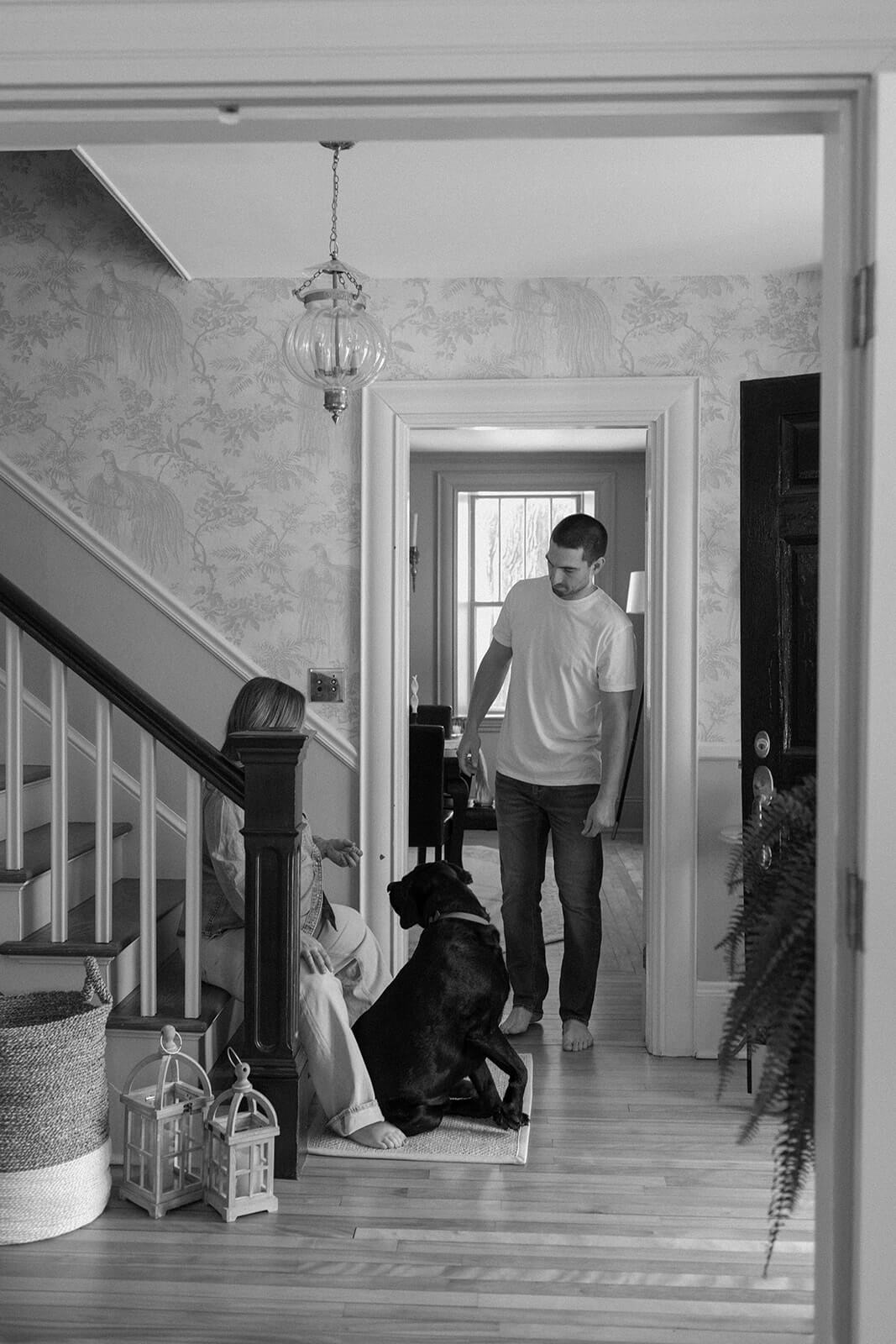 Black-and-white maternity photoshoot at home showing an expecting father standing with the family dog in the hallway.