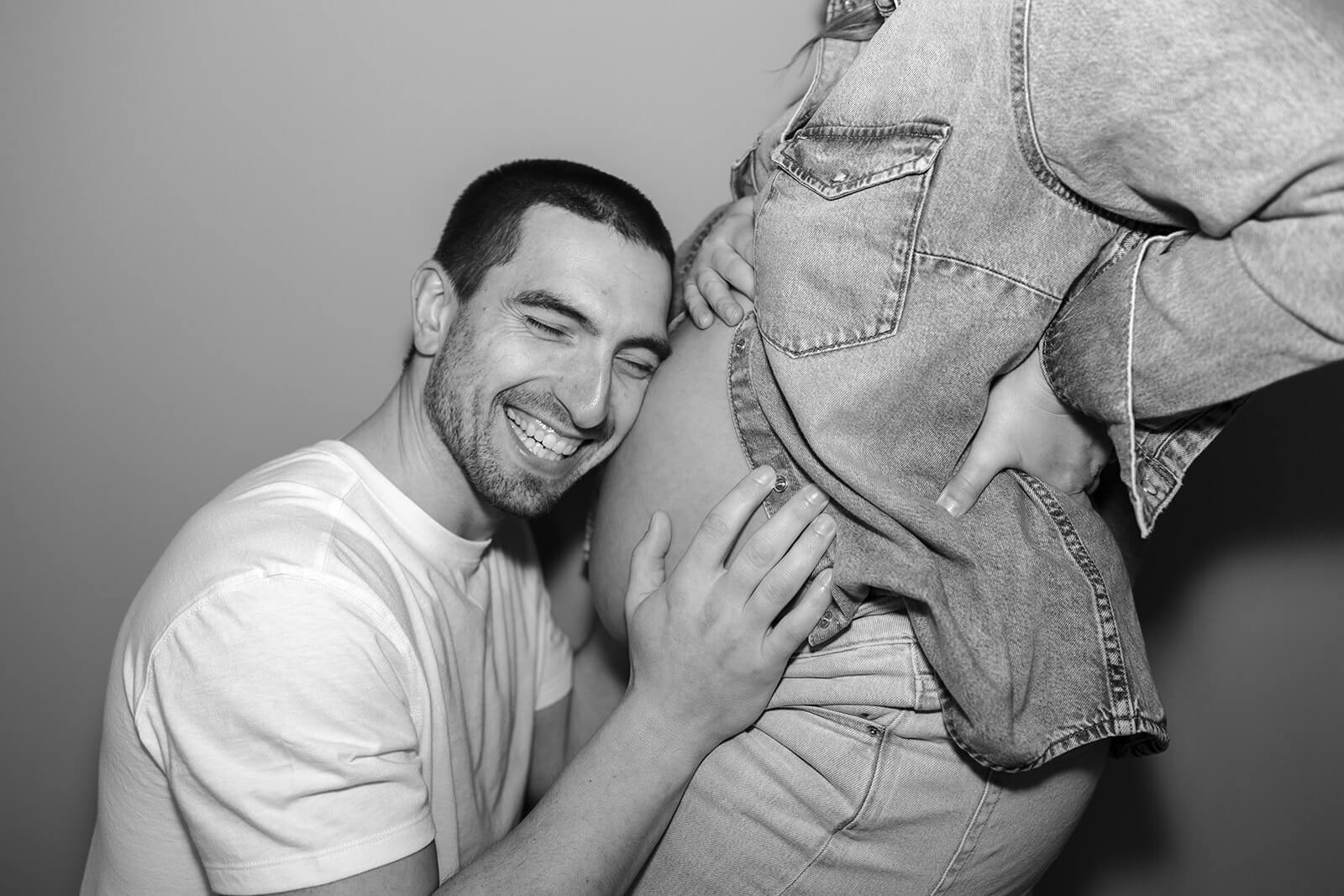 Black-and-white maternity photoshoot at home of an expecting father smiling with his face and hands against his partner’s baby bump.