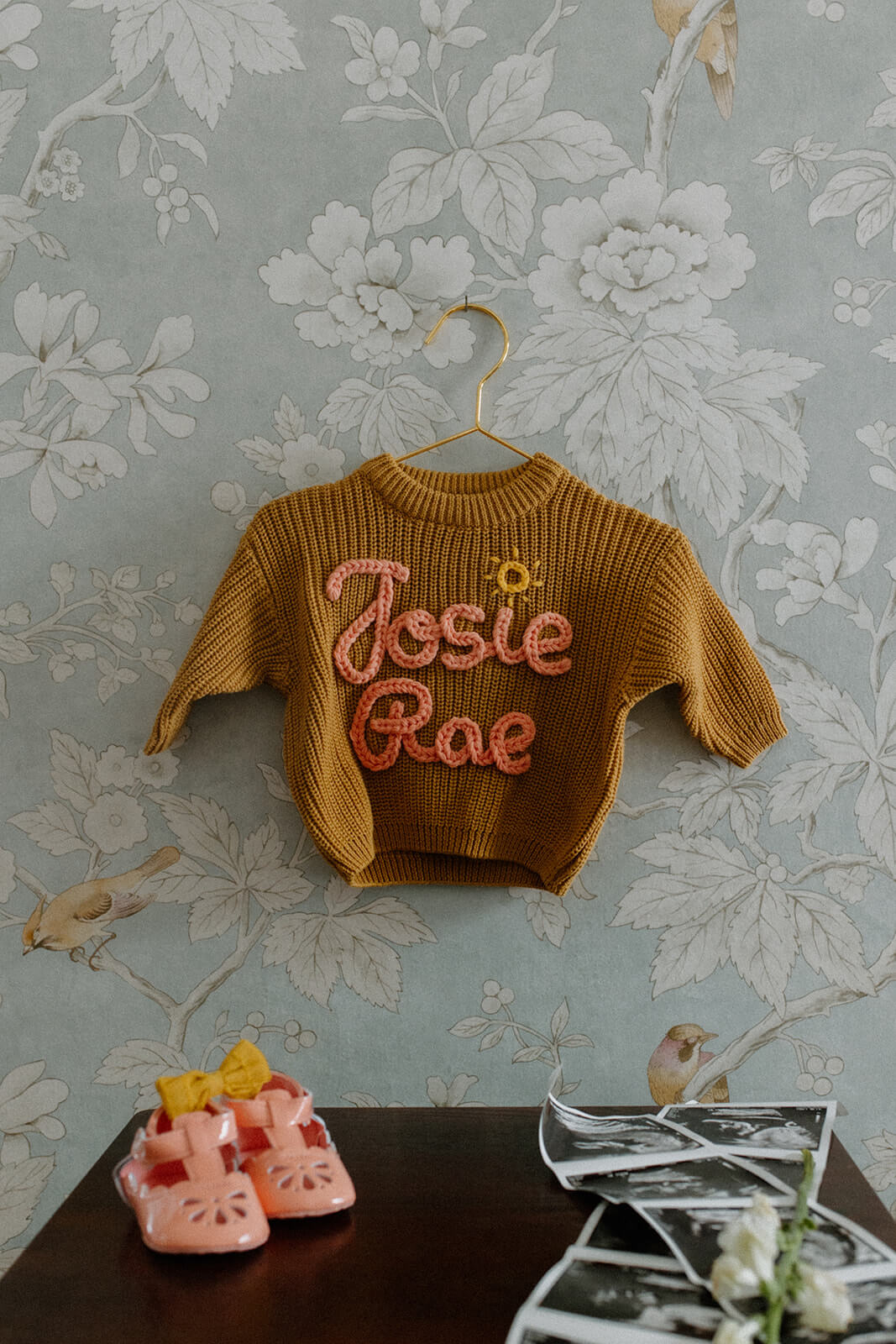 Maternity photoshoot at home detail of a personalized baby sweater hanging above tiny shoes and ultrasound photographs.