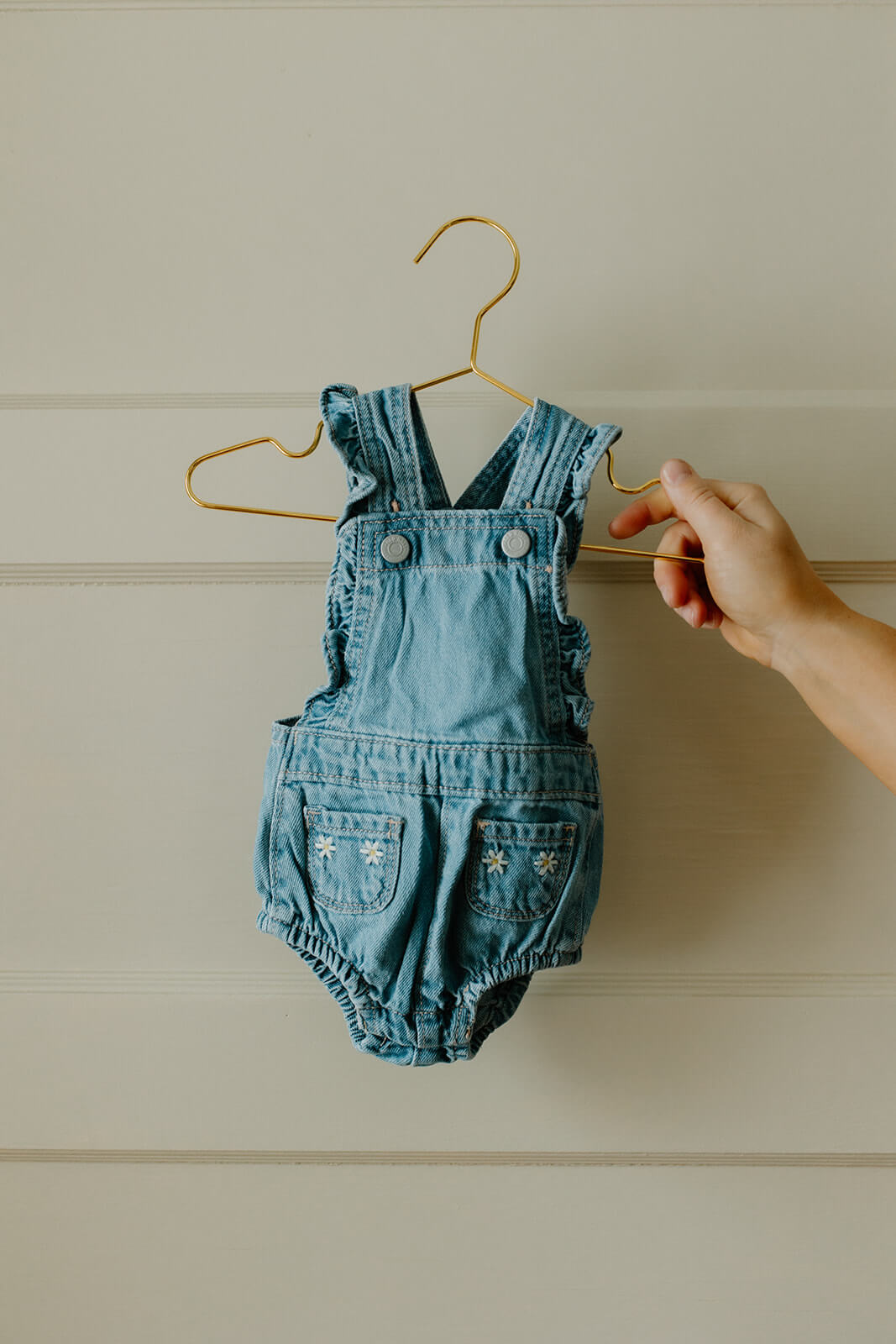 Maternity photoshoot at home detail photo of tiny denim baby overalls hanging from a gold hanger.