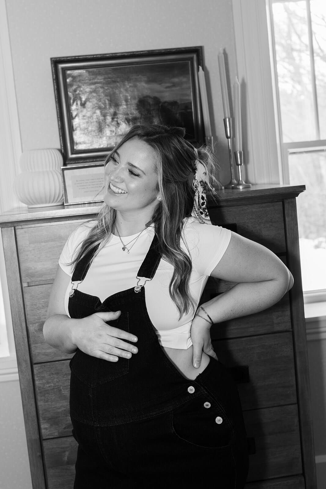 Black-and-white maternity photoshoot at home portrait of an expecting mother in black overalls smiling beside a wooden dresser.