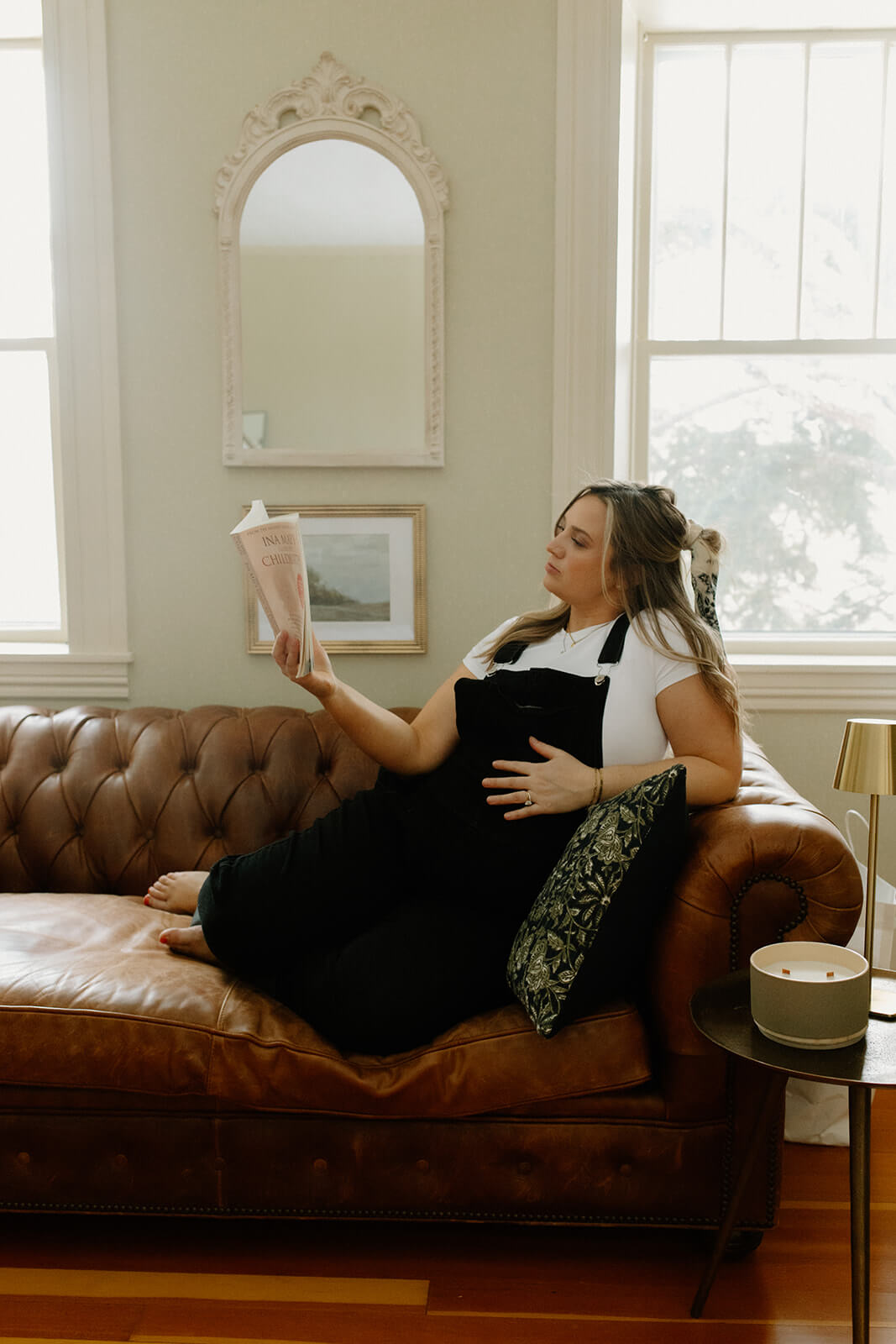 Maternity photoshoot at home of an expecting mother relaxing on a leather couch while reading with one hand resting on her belly.