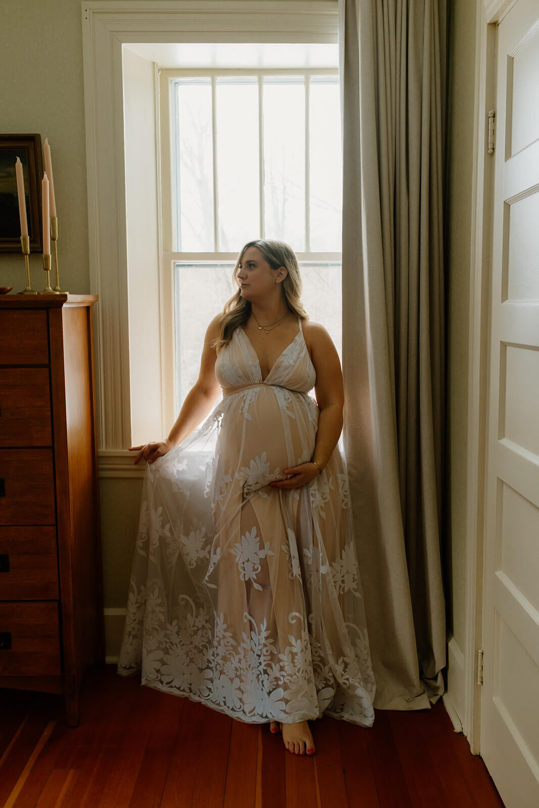 Maternity photoshoot at home portrait of an expecting mother in a sheer floral maternity dress standing beside a window.