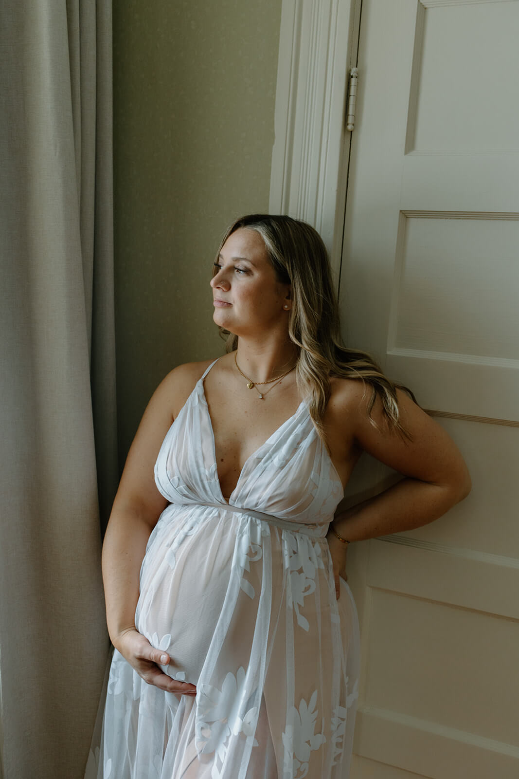Maternity photoshoot at home portrait of an expecting mother standing by a window with one hand supporting her belly.