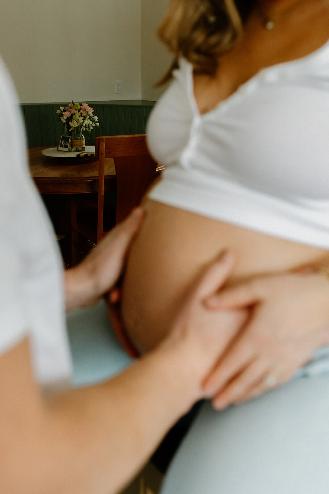 Soft close-up from a maternity photoshoot at home showing an expecting mother’s belly with her partner’s hand resting against it.
