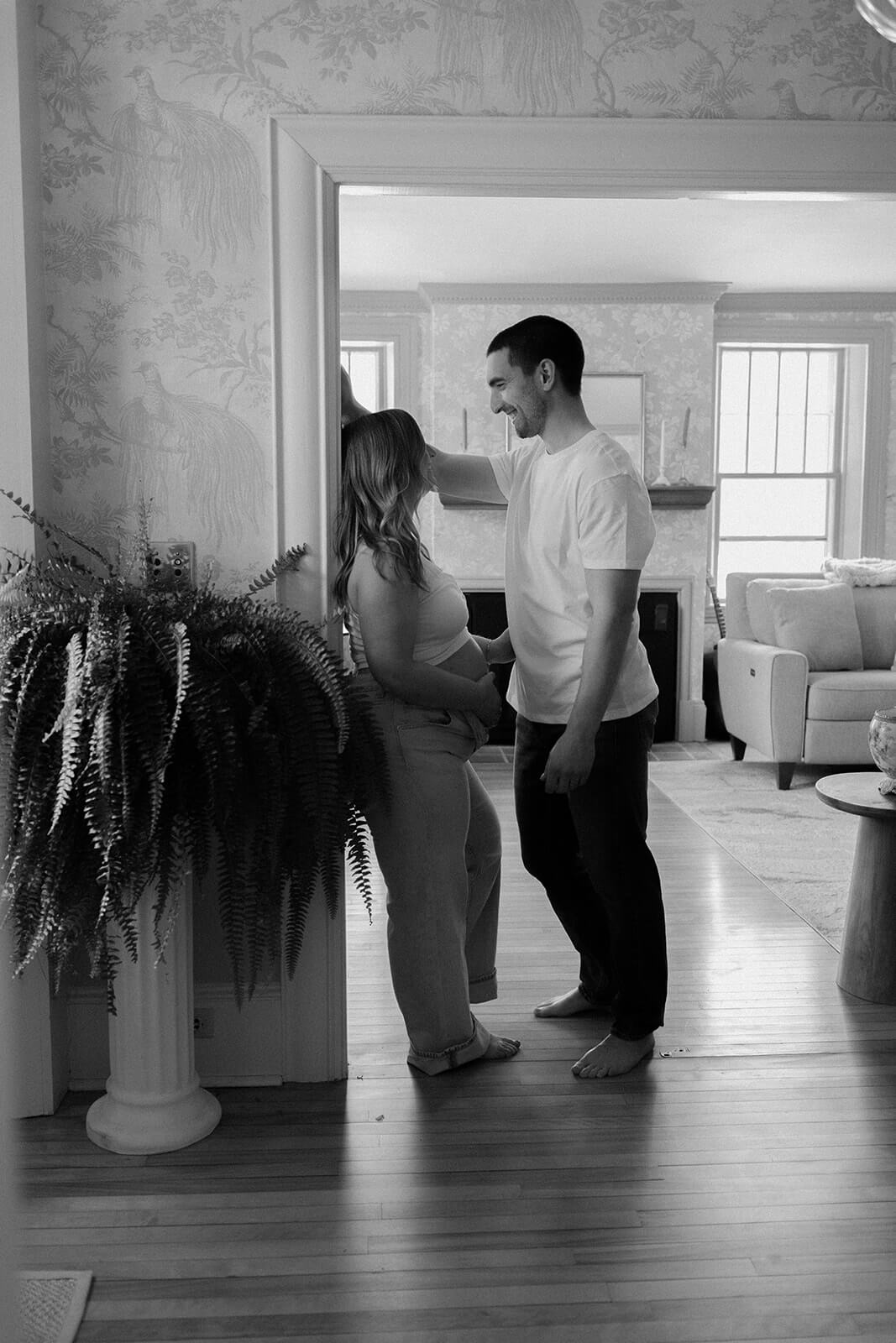 Black-and-white maternity photoshoot at home of an expecting couple smiling at each other in a doorway.