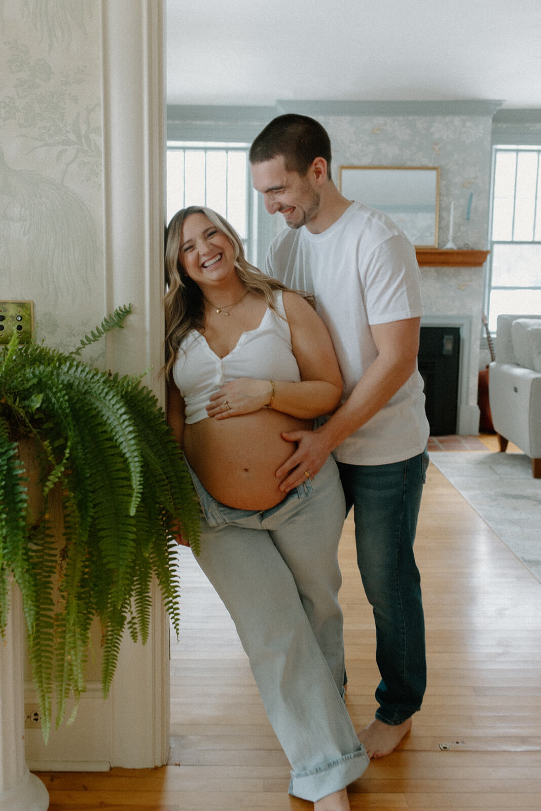 Maternity photoshoot at home of an expecting couple laughing together in a doorway with their hands around her belly.